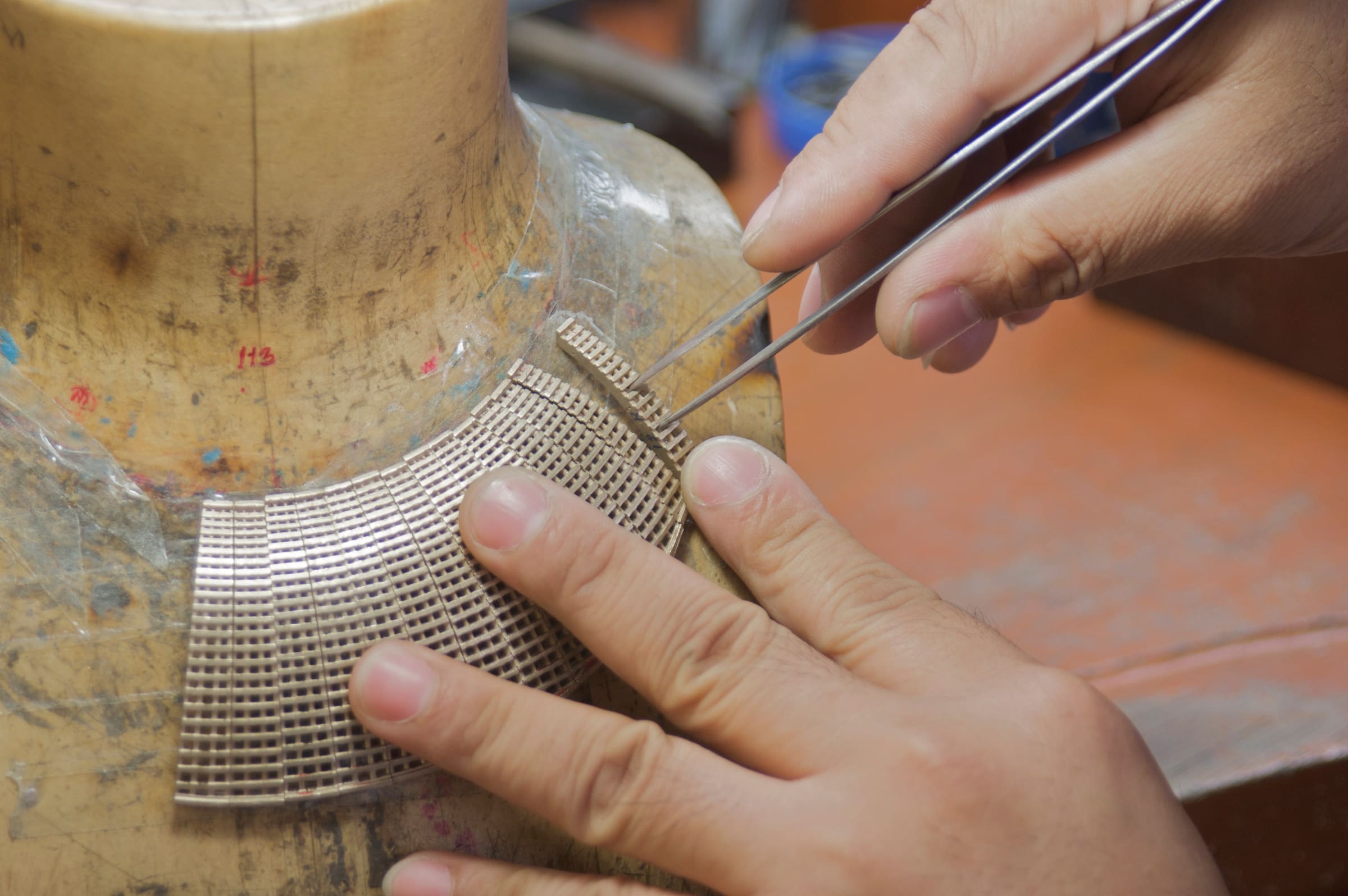 A collar of pierced metal mesh is assembled panel by panel on a wooden neck form.