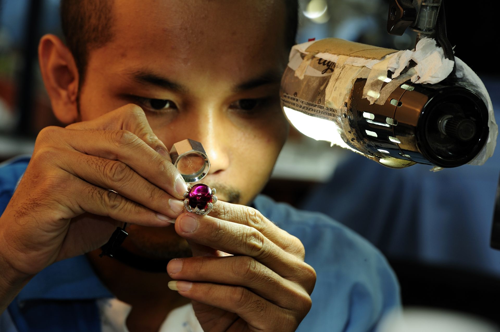 A craftsman examines a cabochon ruby ring set with diamonds through a loupe under the bench lamp.